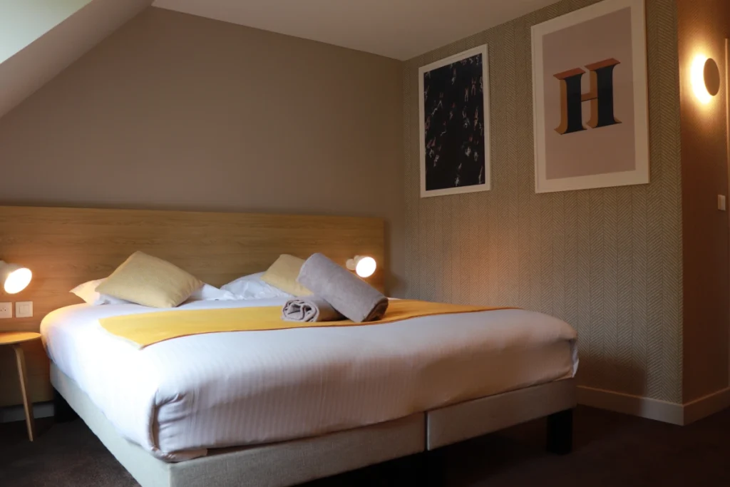 Chambre d'hôtel double avec lit confortable, draps blancs, coussins jaunes, tête de lit en bois, lampes de chevet, décoration murale et éclairage ambiant chaleureux.